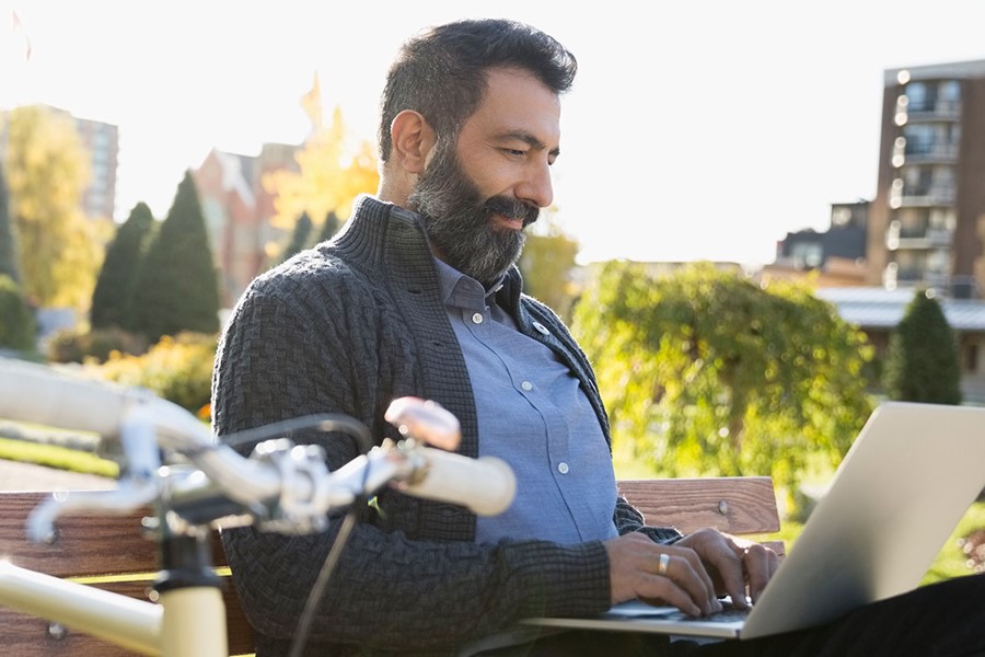 Commuter With Bicycle Using Laptop On Park Bench