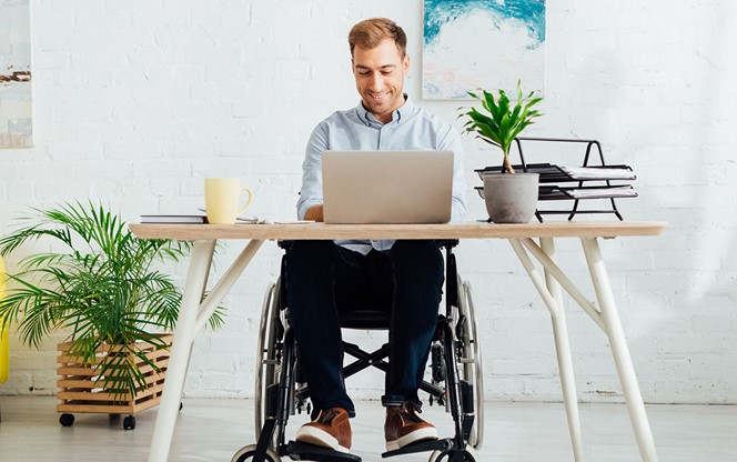 Smiling Freelancer In Wheelchair Using Laptop At Desk In Living Room