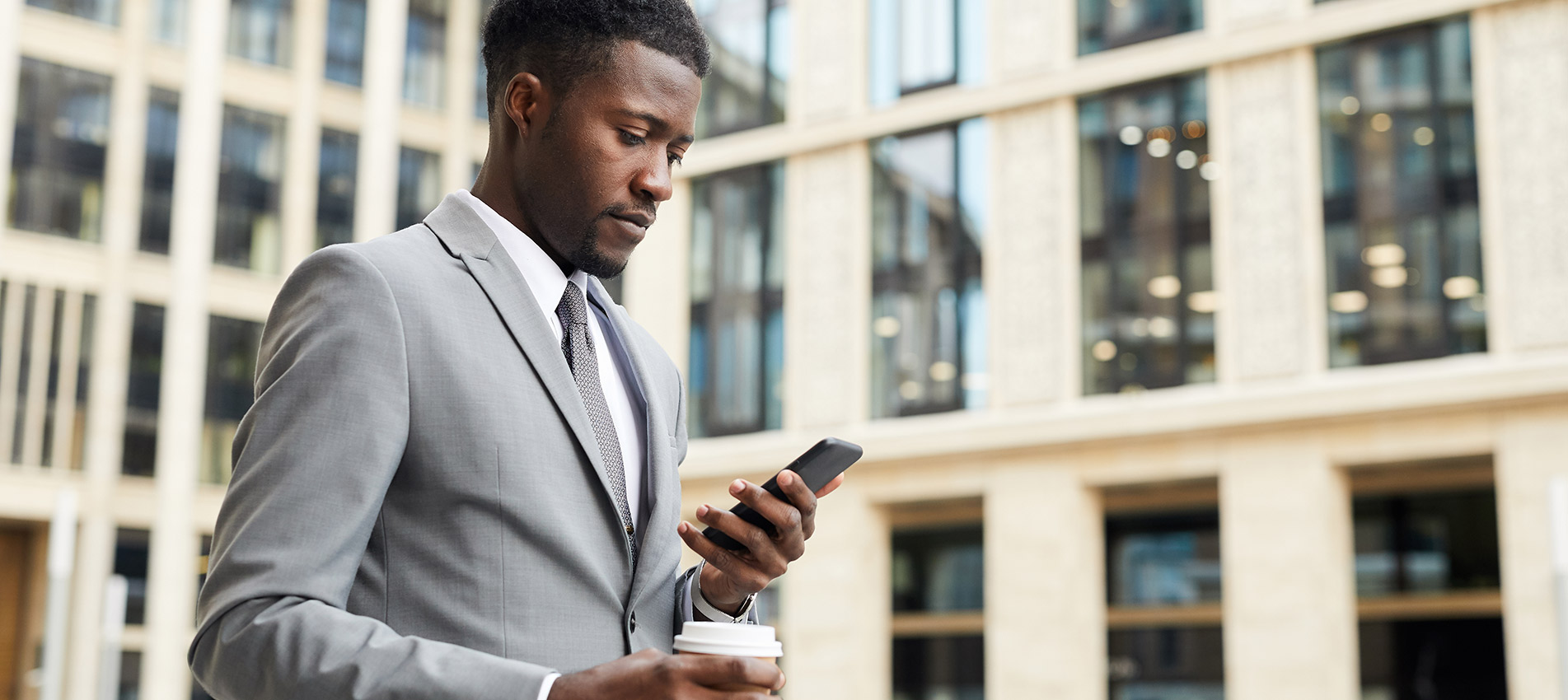 Serious African Businessman In Suit Drinking Coffee And Reading A Message On His Mobile Phone While Walking In The City