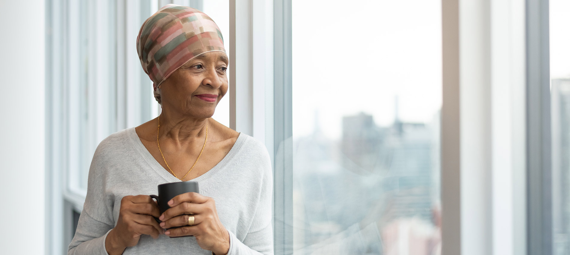 A Black Senior Woman With Cancer Is Wearing A Scarf On Her Head Standing Indoors Looking Out A Bay Of Windows At The City Below