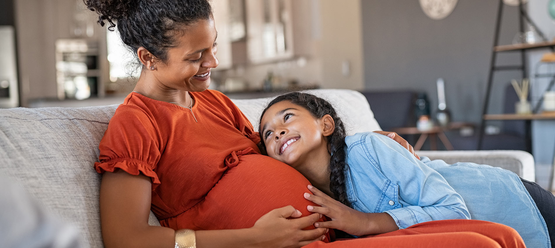 Happy Mixed Race Daughter Hugging Belly Of Her Expecting Mother While Relaxing On Couch At Home