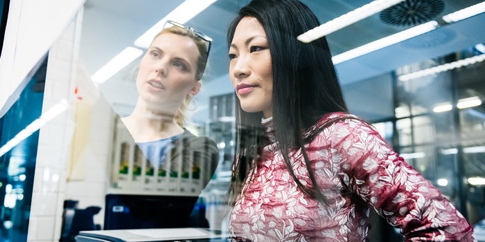 Two Female Engineers Discussing A Project In An Industrial Office While Looking At Documents Pinned On A Glass Wall