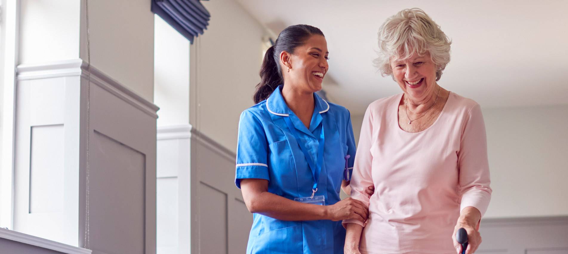 Senior Woman At Home Using Walking Stick Being Helped By Female Care Worker In Uniform