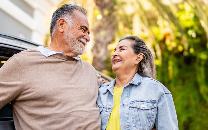 Happy Hispanic Senior Couple Standing Next To A Car At New House