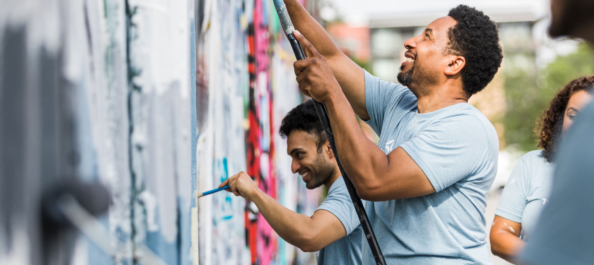Coworkers Smiling While They Paint A Wall At A Community Event