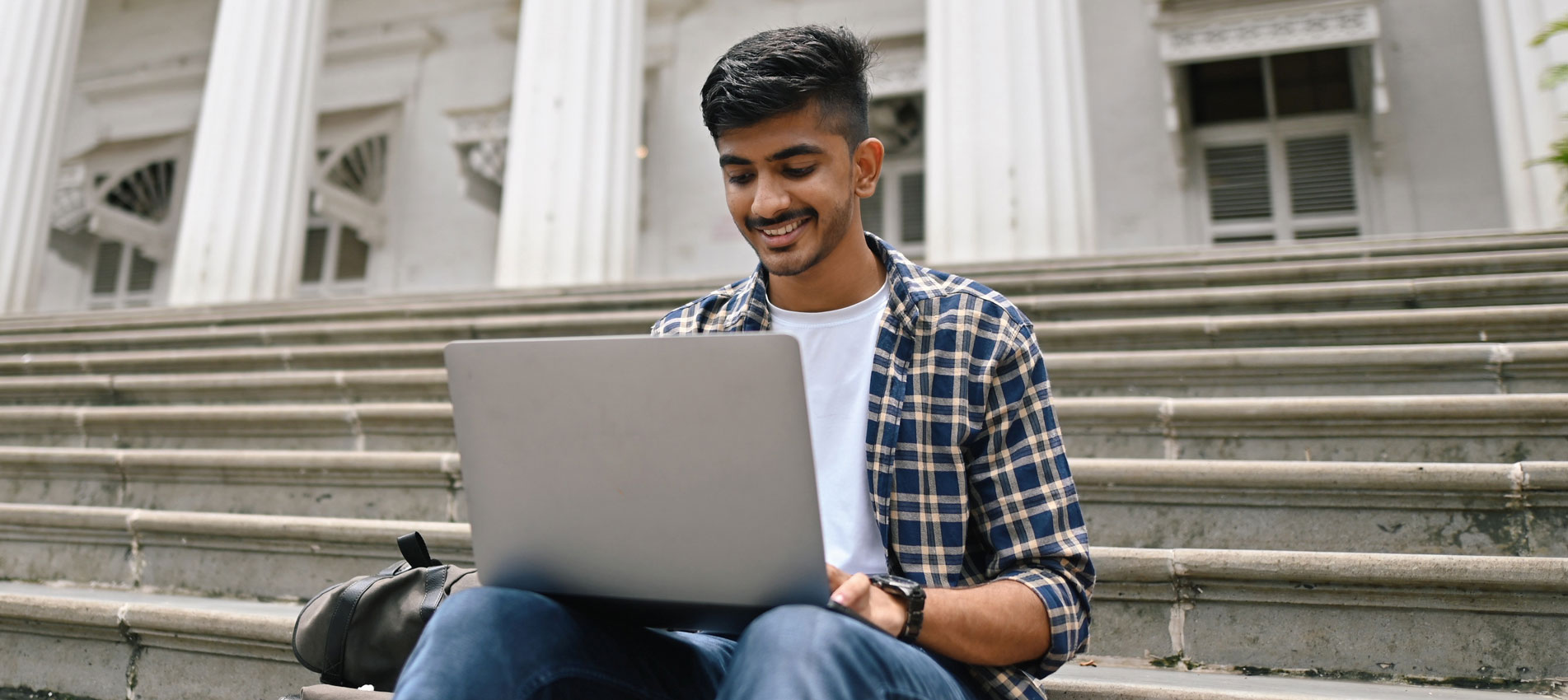 University Student Sitting Outside On Steps And Using Laptop
