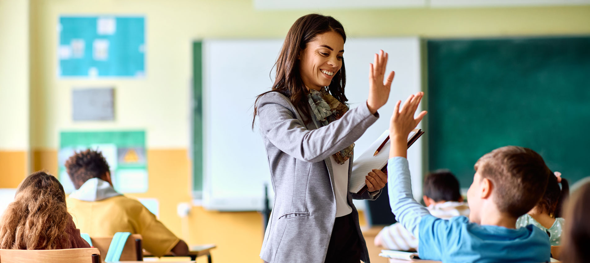 Happy Hispanic Female Elementary School Teacher Giving High Five To Her Student In The Classroom