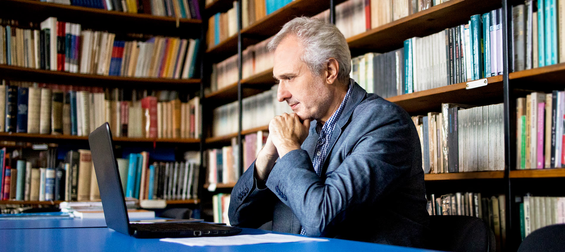 University Senior Professor In Library Looks At Laptop