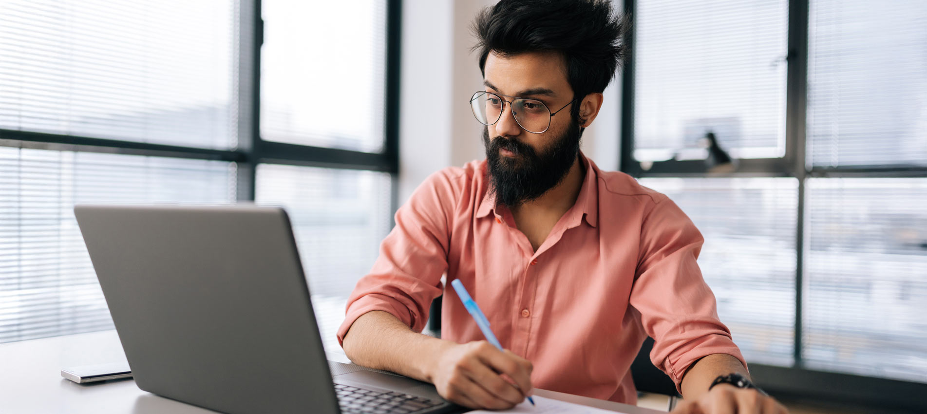 Indian Businessman In Glasses Using Laptop Writing Notes Siting At Table