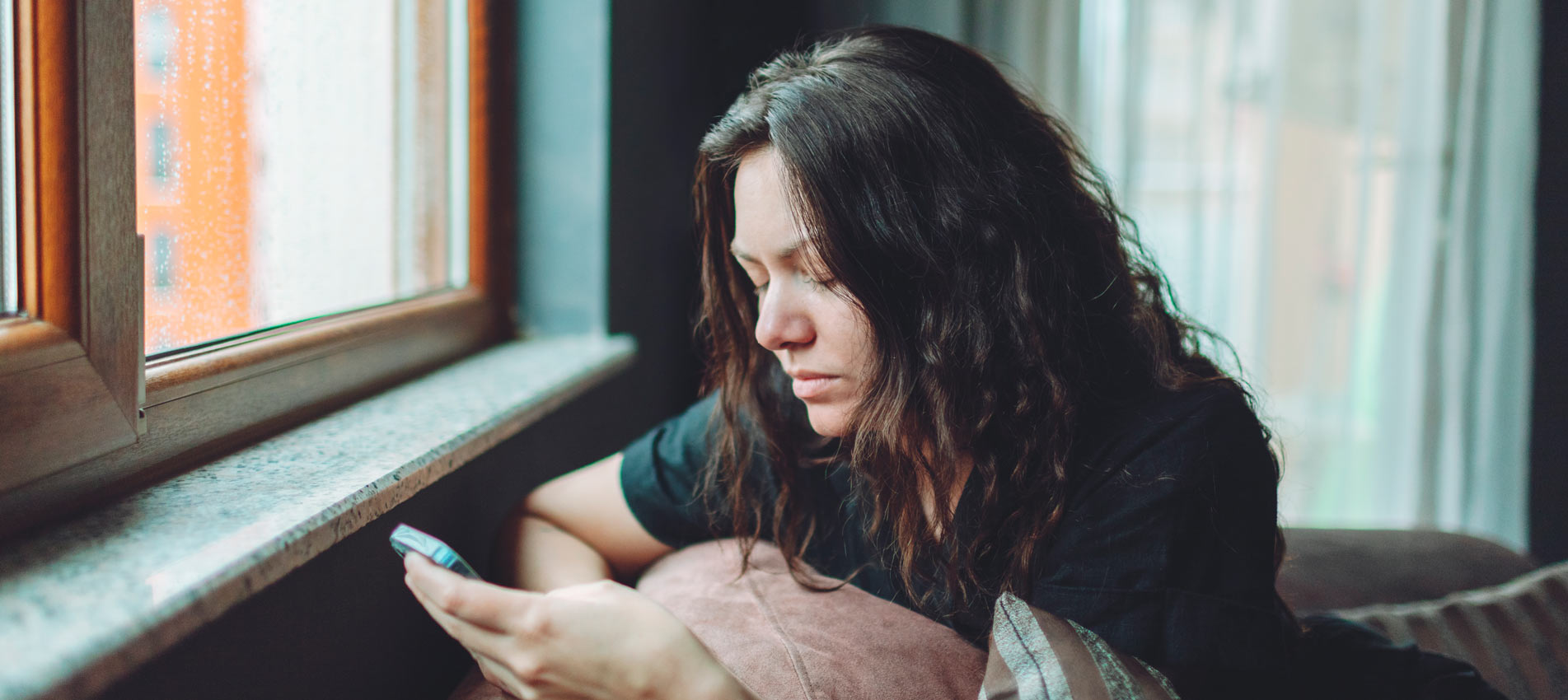 Depressed Woman Sitting On Sofa At Home With Mobile Phone